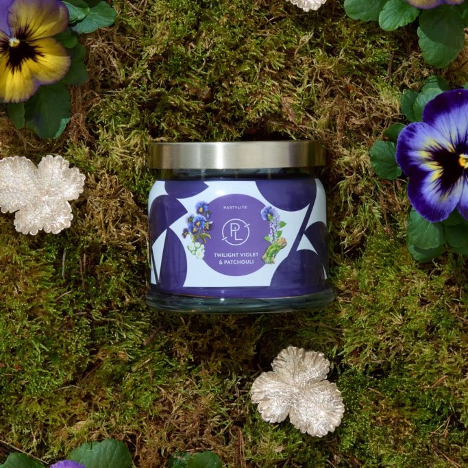 Scented candle in a glass jar surrounded by green moss and flowers.